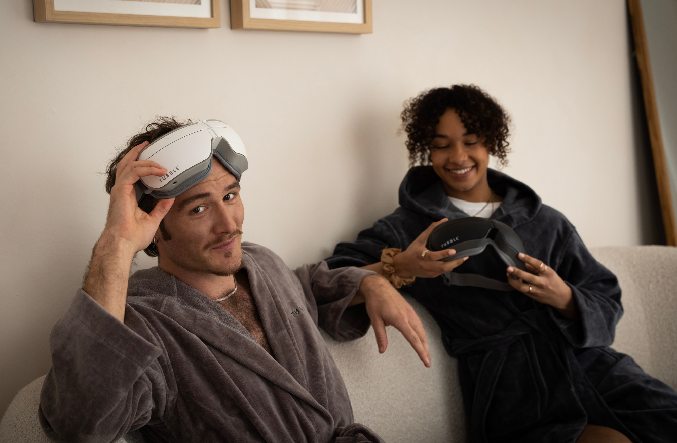 Man and woman relaxing on a sofa in robes while holding Tubble eye massagers, smiling and preparing to use the devices