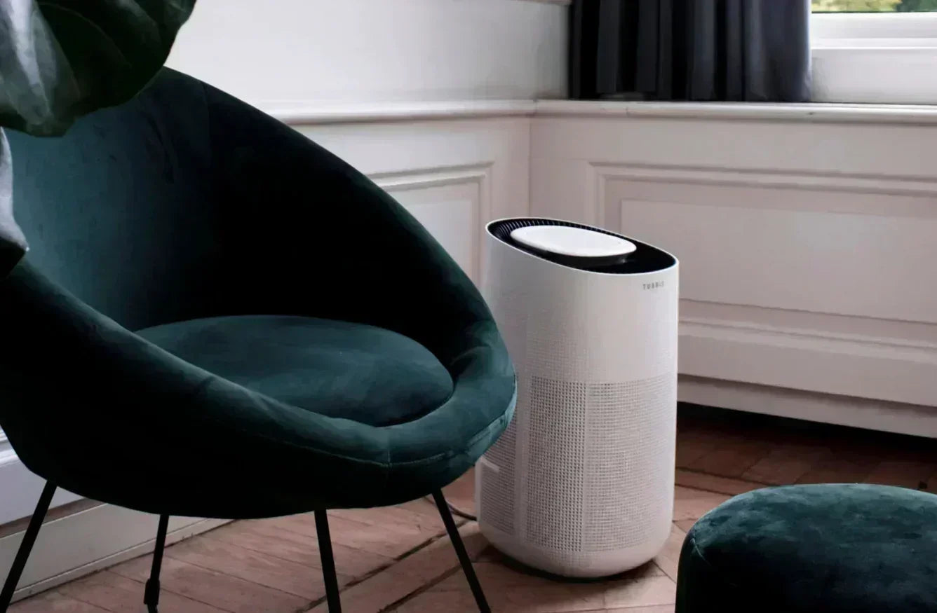 Air purifier on a wooden floor next to a green chair in a room with white walls.
