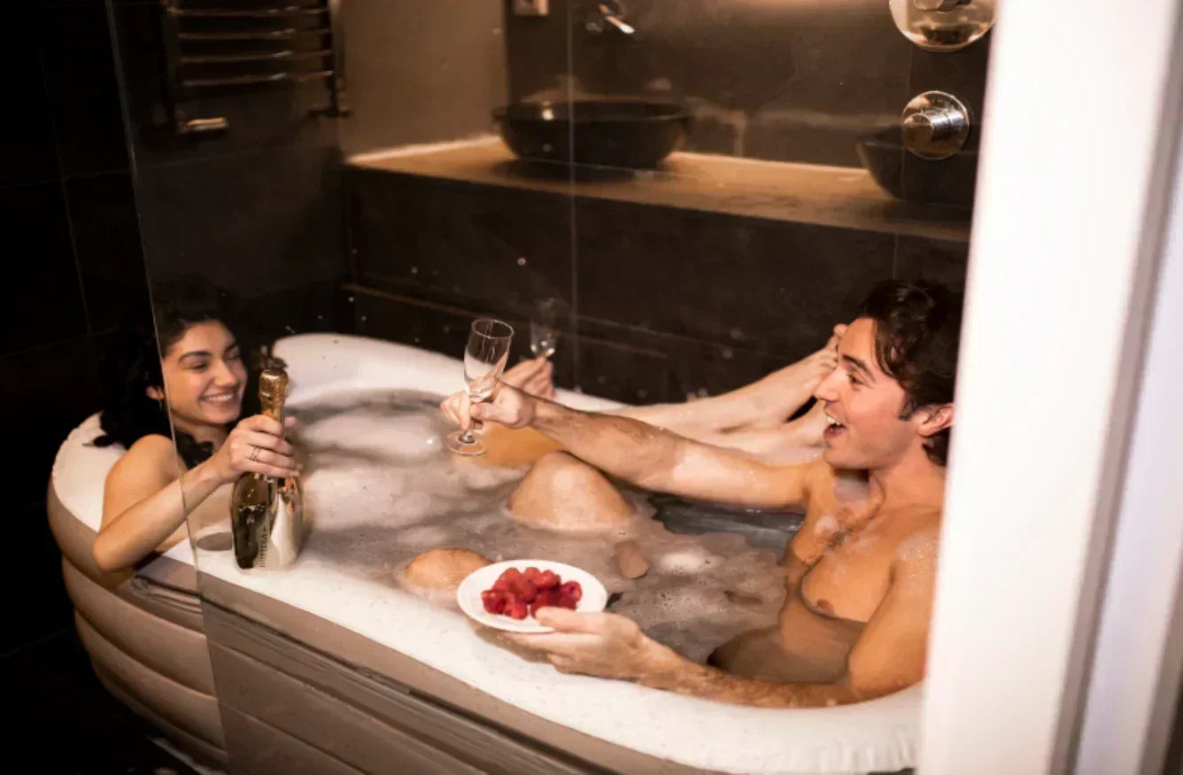 Two people in a bubble bath with a bottle of champagne and a plate of raspberries.