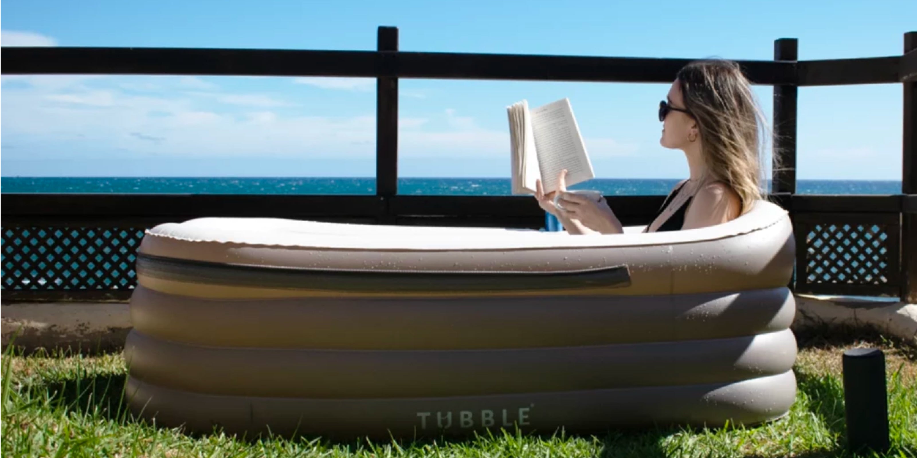 Woman relaxing in a Tubble inflatable bathtub outdoors by the sea, reading a book on a sunny day.