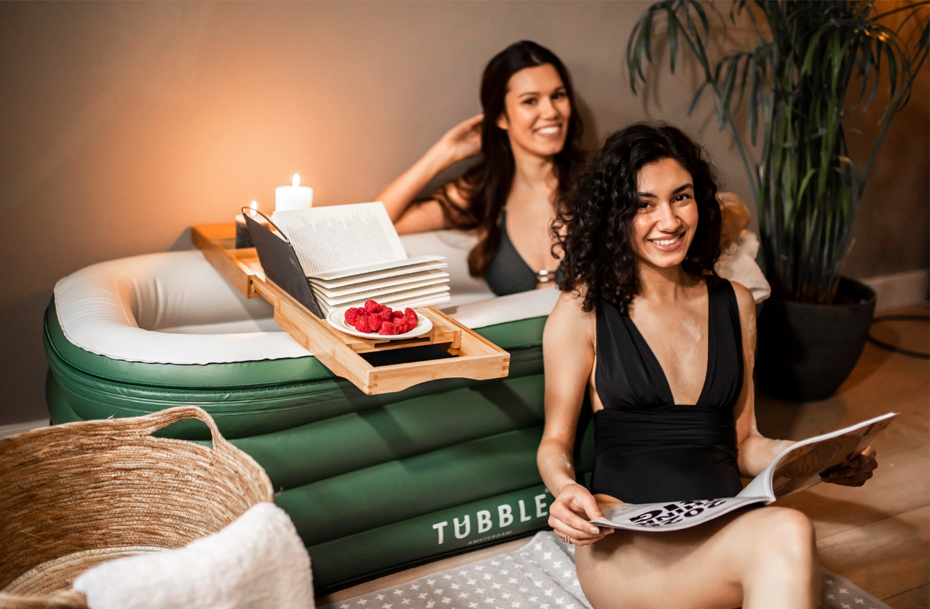Two women relaxing in a green Tubble inflatable bathtub with a bath tray holding candles, a book, and raspberries.