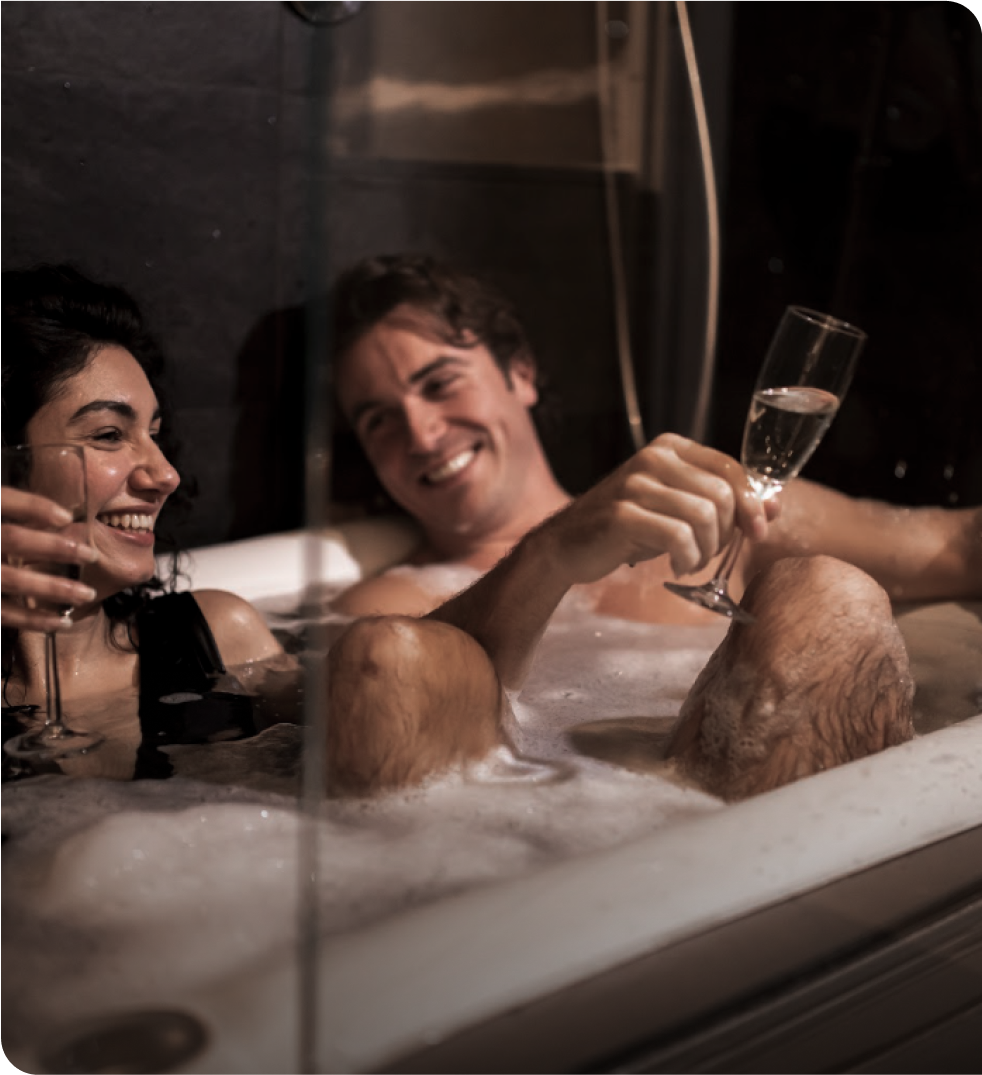 Romantic couple relaxing together in a warm bubble bath, smiling and enjoying champagne in a cozy spa-like bathroom setting, promoting luxury home wellness and intimate relaxation experiences.